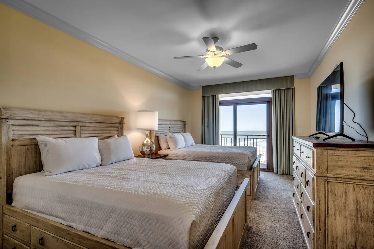 A spacious bedroom shows two large beds set beside a window with a scenic view, along with a television and dresser. The room exudes a calm, welcoming mood, suitable for families or groups wanting to share a space without sacrificing comfort or privacy.
