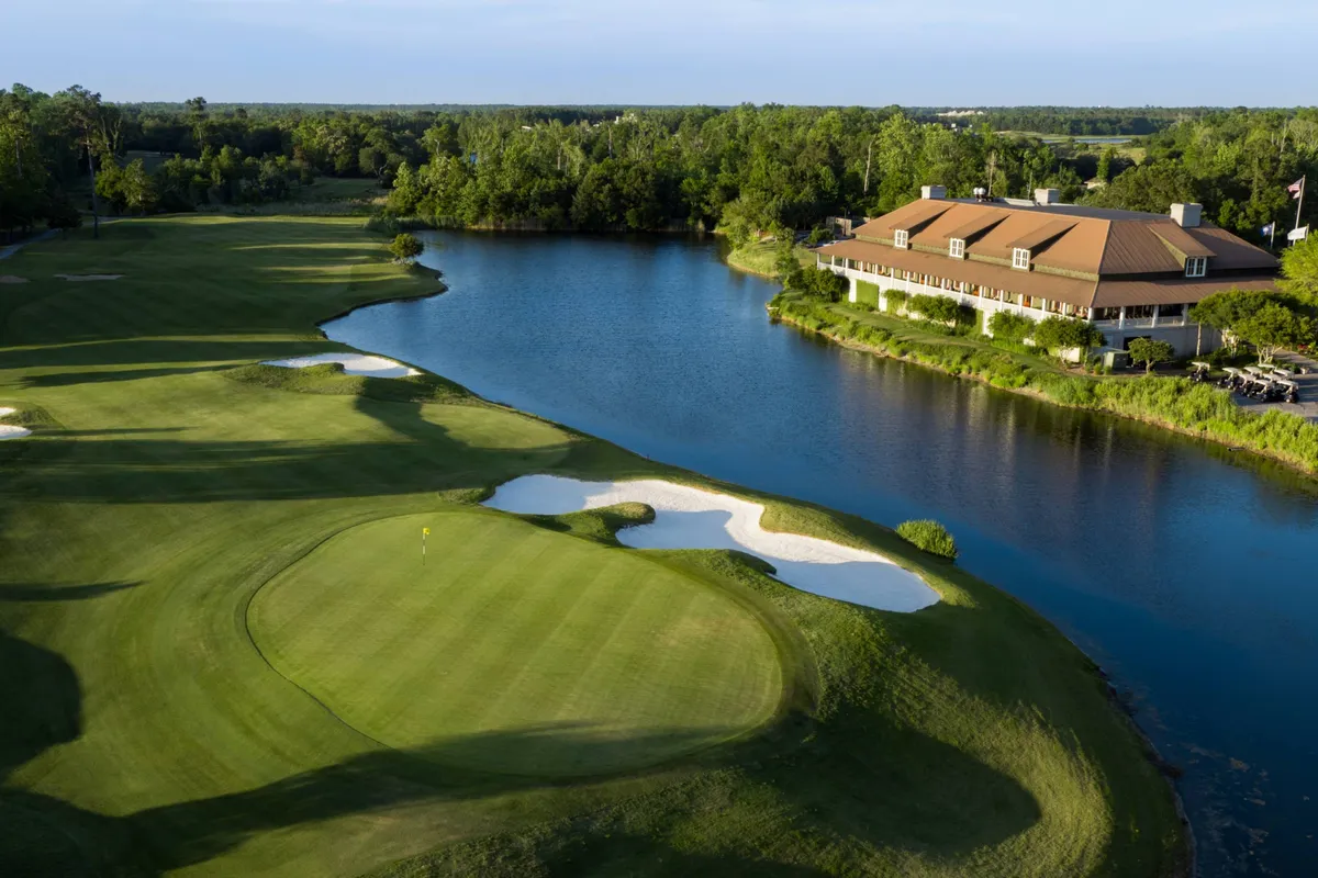 A lush golf course stretches alongside a tranquil lake, with a clubhouse in the distance and picturesque sand traps dotting the landscape. The peaceful setting is perfect for a day of golfing, aligning with the mention of complimentary access to Azalea Sands Golf Club.