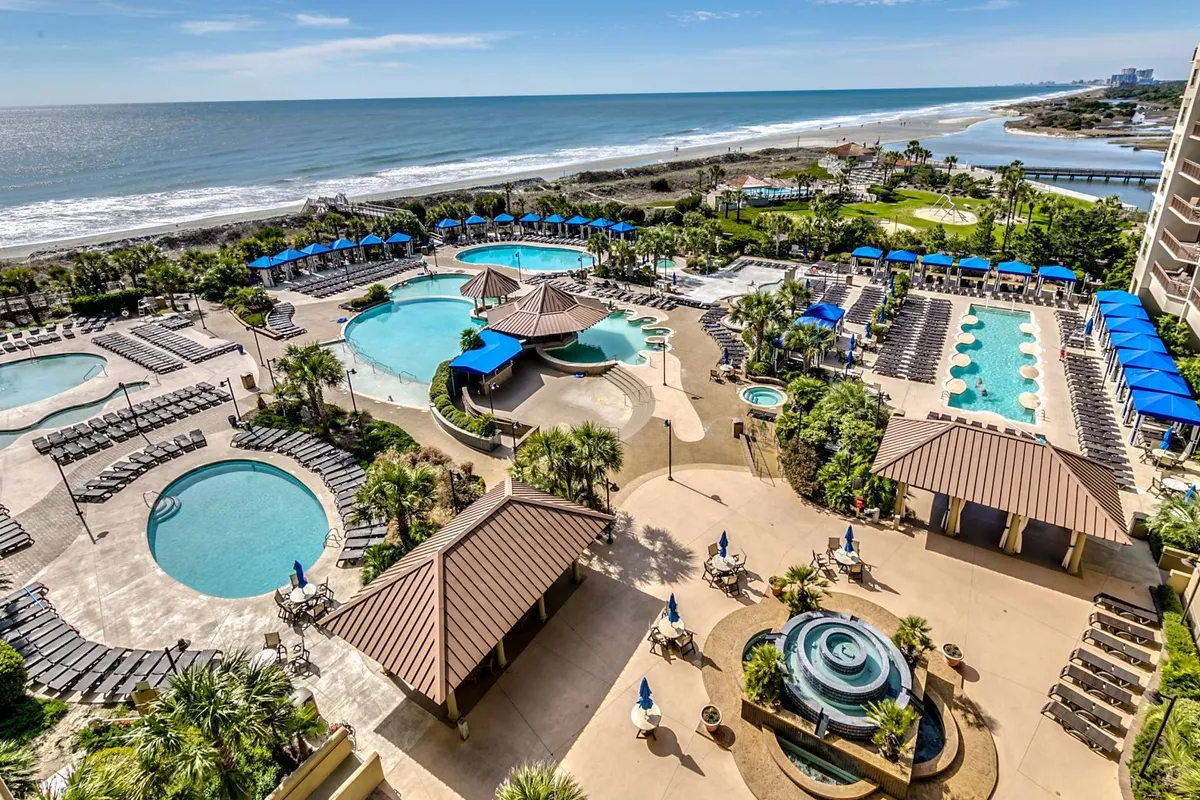 A sweeping exterior view of the resort’s pool complex, featuring multiple swimming pools surrounded by lush palm trees, beachfront vistas, and deck chairs shaded by umbrellas. This scene captures the lively, leisurely atmosphere of the resort, inviting residents to swim, sunbathe, or relax by the water at any time of day.