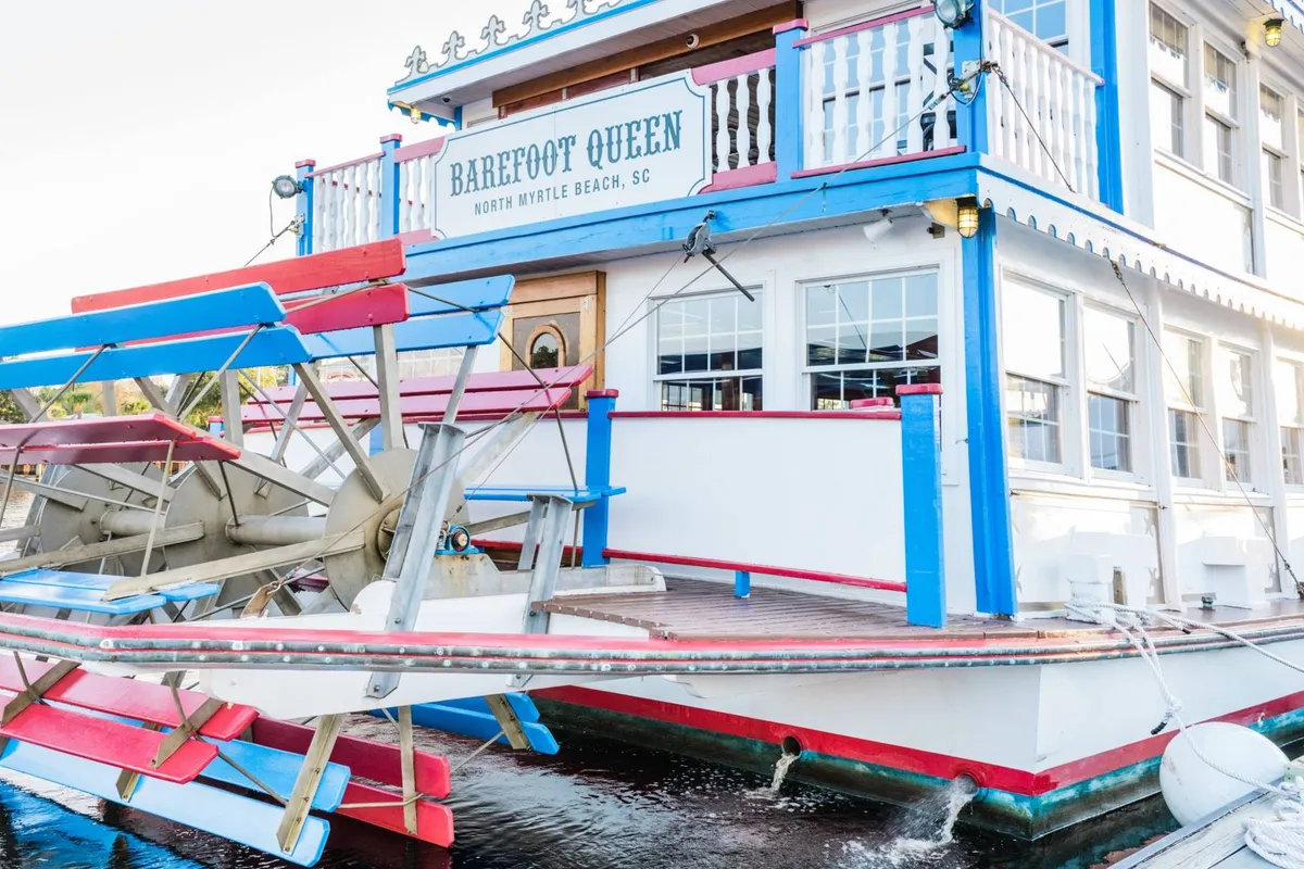 A colorful paddle wheel riverboat is docked, its distinctive exterior gleaming in the light. The scene suggests festive social gatherings, sunset cruises, and unique dining experiences on the water—mirroring the Barefoot Queen Riverboat Dinner Cruise included for guests.