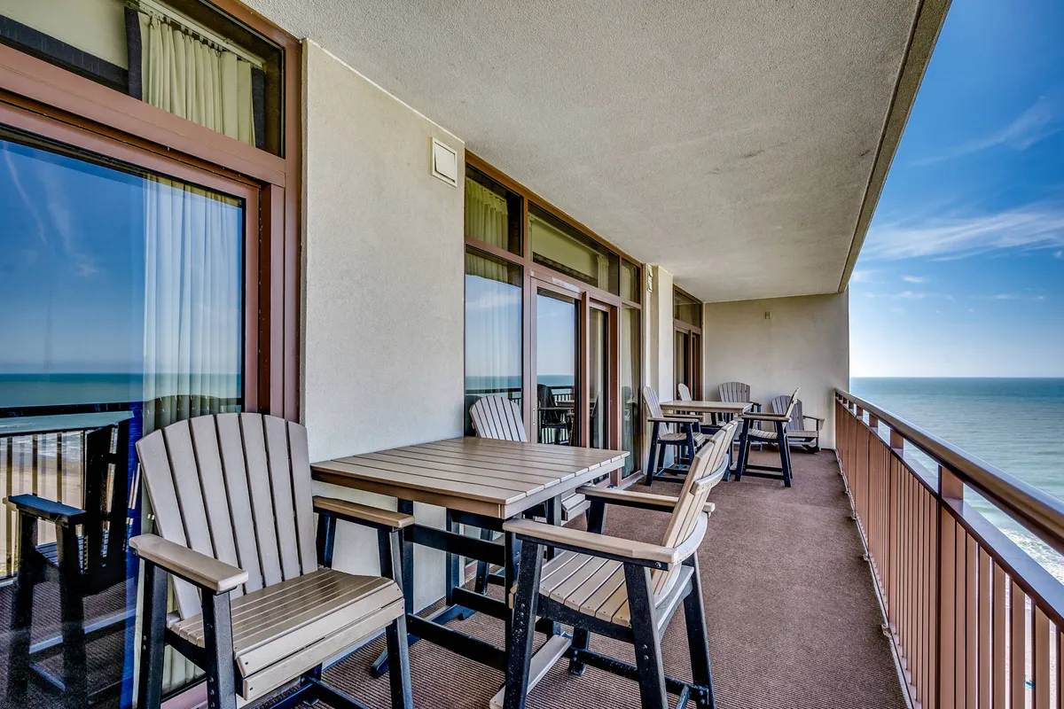A private balcony with comfortable seating overlooks the ocean. A table is set for leisure activities like morning coffee or reading, and the view stretches out across shimmering water, capturing the tranquil beachfront atmosphere.