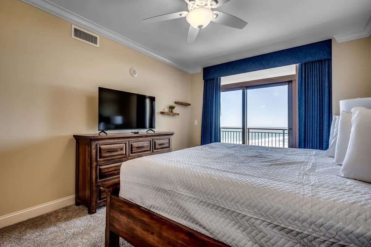 A serene bedroom with a plush bed, a large window offering views of the outdoors, a television, and a dresser. The setup emphasizes relaxation, privacy, and the luxury of waking up to ocean views.
