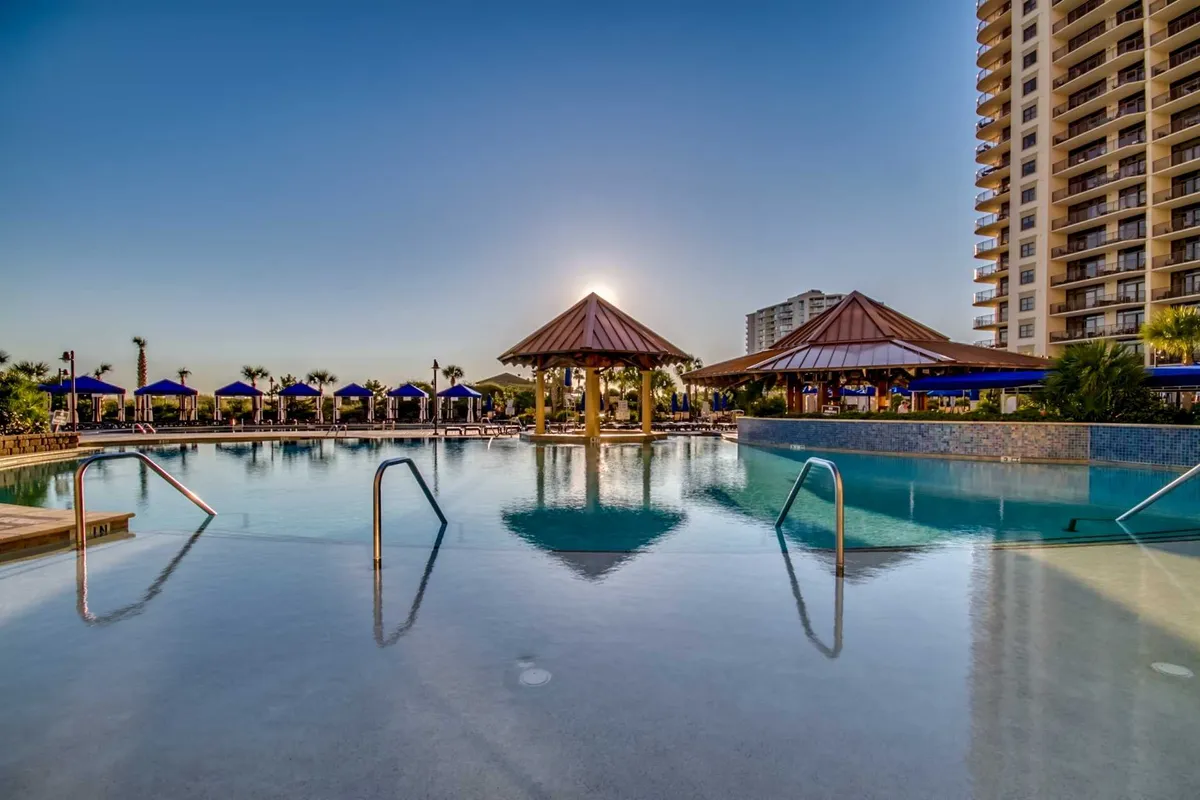 A sprawling resort pool complex at sunset, with thatched-roof cabanas, a shimmering swimming pool, and guests lounging poolside. The golden light creates an inviting and serene ambiance, ideal for both families and adults.