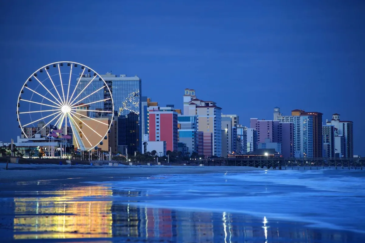 A ferris wheel stands tall against a backdrop of colorful seaside buildings, its lights reflecting off the ocean. The beachside promenade invites guests to explore local attractions and soak up the playful coastal energy.